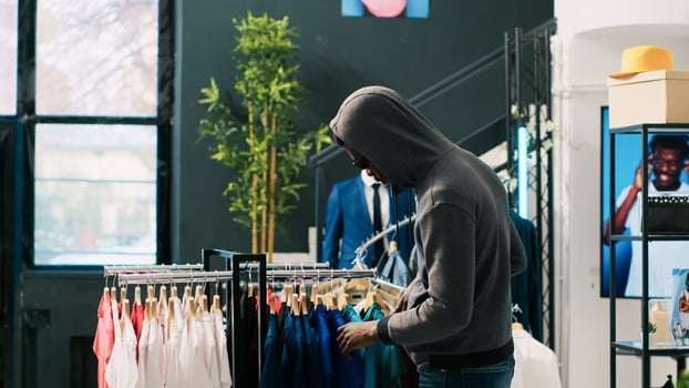 African american thief robbing clothing store, analyzing racks with stylish clothes. Robber trying to steal fashionable merchandise, wearing sunglasses and hood not to be recognized photo