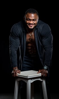 Smiling cheerful young black man in black shirt leaned with hands on chair. Unbuttoned shirt shows perfect muscular body. Dark background. photo