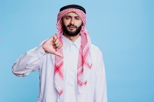 Muslim man dressed in traditional arabian clothes showing thumb down studio portrait. Arab in thobe and ghutra showcasing disagreement and looking at camera with serious expression photo