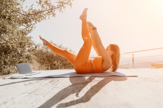 Fitness woman sea. Happy middle aged woman in orange sportswear exercises morning outdoors on yoga mat with laptop in park over ocean beach. Female fitness pilates yoga routine. Healthy lifestyle. photo