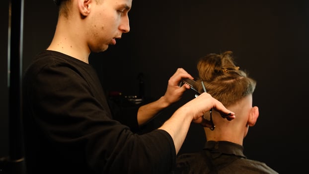 Barber use Thinning scissors and metal comb on brown wavy hair of young man. Hairdresser service in a modern barbershop in a dark key lightning with warm light back view photo