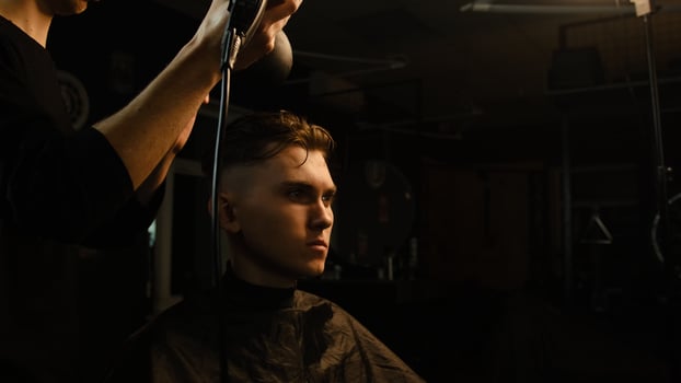 Young barber drying hair of customer with hair dryer in barber shop in a dark key lightning with warm light and correct hair with hand. Hairdresser for man photo