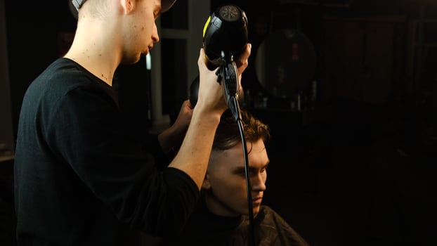 Young barber drying hair of customer with hair dryer in barber shop in a dark key lightning with warm light and correct hair with hand. Hairdresser for man photo