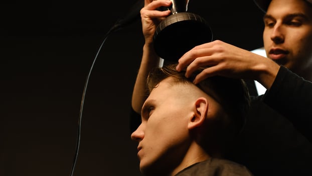 Young barber drying hair of customer with hair dryer in barber shop in a dark key lightning with warm light and correct hair with hand. Hairdresser for man photo