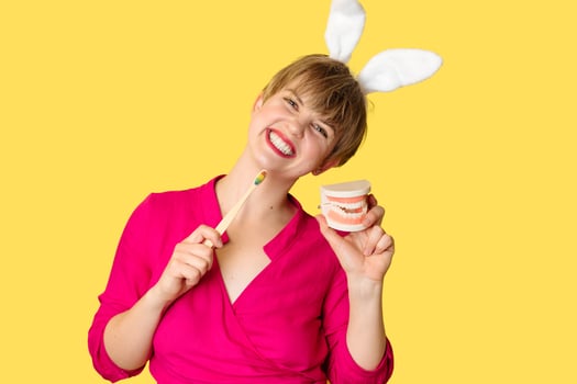 A young woman in bunny ears with an amazing smile with a toothbrush and a jaw model in her hands on a yellow background. photo