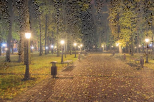 Night rainy park with yellow maple leaves, pavement and lanterns behind wet rainy glass in golden autumn. The concept of bad weather, change of season and leaf fall. Abstract landscape. photo