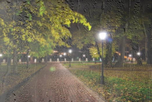 Night rainy park with yellow maple leaves, pavement and lanterns behind wet rainy glass in golden autumn. The concept of bad weather, change of season and leaf fall. Abstract landscape. photo