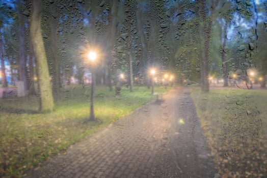 Defocused night park with lanterns behind rainy, wet glass at night. Concept of rainy autumn weather. photo