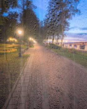Defocused night park with lanterns behind rainy, wet glass at night. Concept of rainy autumn weather. photo
