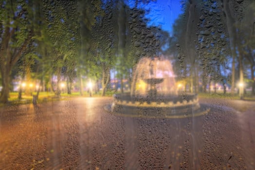Defocused fountain in a night park with lights behind rainy, wet glass at night. Concept of rainy autumn weather. photo