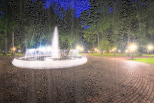 Defocused fountain in a night park with lights behind rainy, wet glass at night. Concept of rainy autumn weather. photo