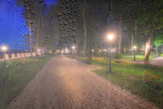Defocused night park with lanterns behind rainy, wet glass at night. Concept of rainy autumn weather. photo