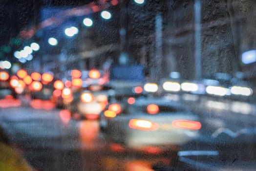 Blurry moving cars with headlights and lanterns reflecting on the wet asphalt in the night city behind rain-covered window. photo