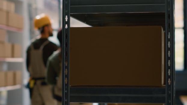 Warehouse employees in blurry backgrounds perform inventory control, keeping quality standards high. Focus shot on cardboard box parcel in storage deposit ready to be shipped photo