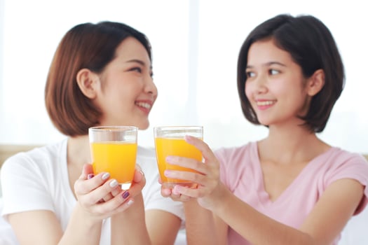Asian young sisters lovely couple on white bed and smiling with hand holding Orange juice glass in bedroom. Holiday and healthy concept. Closed up on Orange juice glass. photo