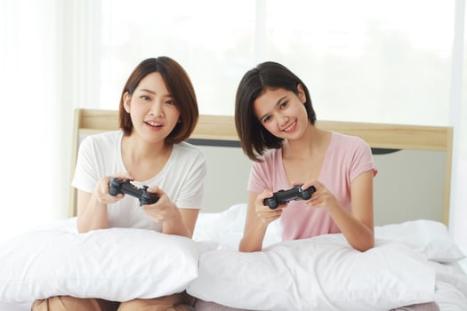 Asian young sisters lovely couple on white bed hand holding joysticks and enjoy with game play station together in bedroom. Hobby and entertainment concept photo