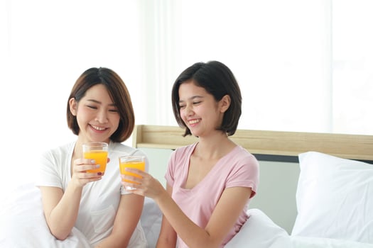 Asian young sisters lovely couple on white bed and smiling with hand holding Orange juice glass in bedroom. Holiday and healthy concept photo