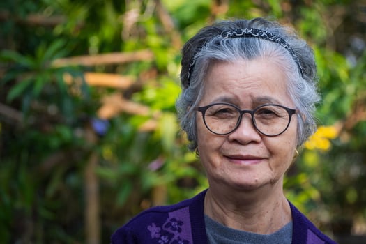 Portrait of a happy elderly woman with short gray hair, wearing glasses, smiling, and looking at the camera while standing in a garden. Space for text. Concept of aged people and relaxation photo