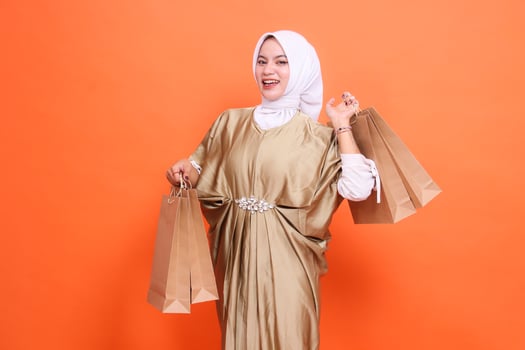 expression of a cheerful young indonesia woman carrying a paper bag containing groceries wearing a kaftan Muslim dress with a hijab, isolated on an orange background. concept transaction, promotion photo