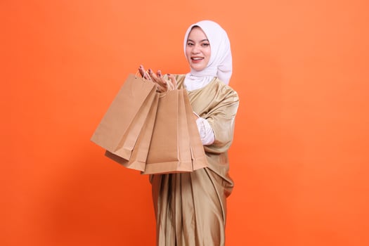Cheerful Asian woman with both hands carrying a brown paper bag containing groceries in front of her wearing a kaftan Muslim dress with a hijab, isolated on an orange background. fashion transaction photo