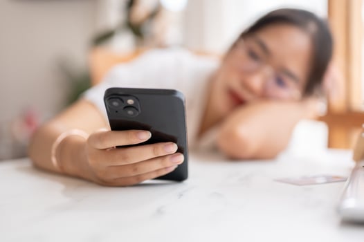 A tired, bored Asian woman leaning on the table and holding her smartphone, waiting for the messages photo