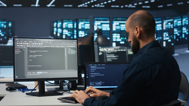 Developer typing code, working in network security data center facility, ensuring optimal performance. IT professional using desktop PC to monitor energy consumption across blade server clusters photo