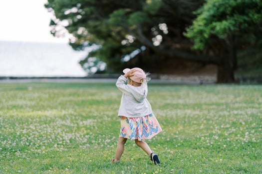 Little girl walks on a green lawn holding a cap on her head. Back view photo