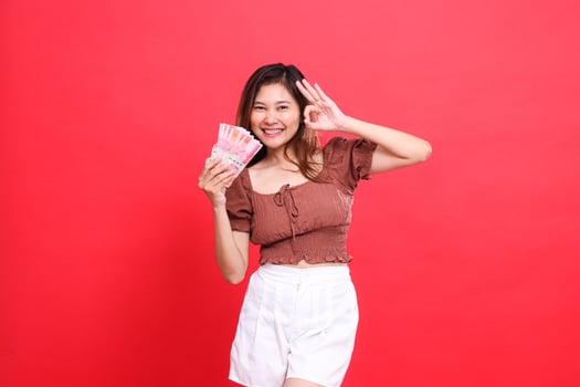 gesture of a cheerful indonesia woman with a zero sign cool, okay while holding rupiah currency, wearing a brown blouse with a red background. for transaction, technology and advertising concepts photo