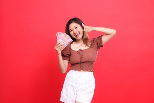 Cheerful Asian woman gestures with a thumbs up sign okay good while holding rupiah currency wearing a brown blouse with a red background. for transaction, technology and advertising concepts photo