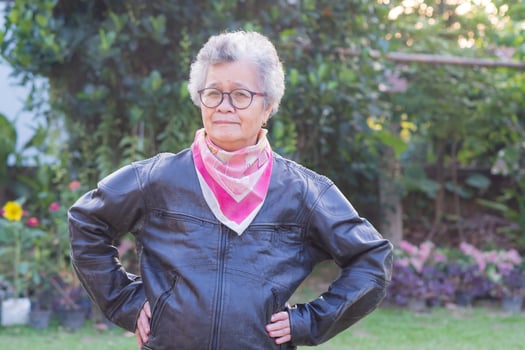An elderly Asian woman with short gray hair wearing a black leather jacket and colorful scarf, standing and arms crossed on light orange background. Classic vintage biker. People and vintage concept photo