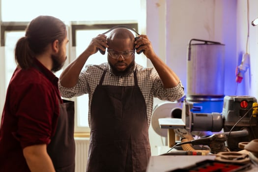 Master in carpentry shop instructing BIPOC worker to wear protection gear before assembling furniture. Cabinetmaker making sure apprentice uses safety earmuffs and glasses before starting work photo
