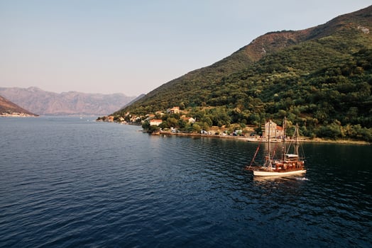 Wooden sailboat sails on the sea along the green mountainous coast. Drone photo