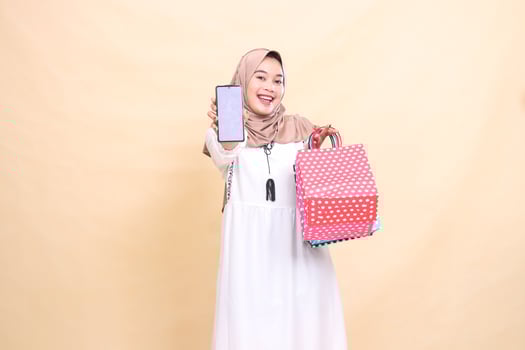 a young Asian Muslim woman in a happy hijab displays a cellphone screen and carries a paperbag shopping bag. for advertising, technology, Eid and Ramadan photo