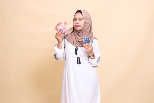 a beautiful Asian Muslim girl in a hijab frowns carrying rupiah notes and a debit credit card for payment. used for advertising, technology, Eid and Ramadan photo