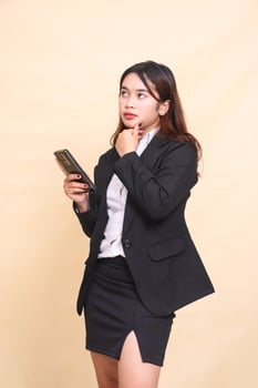 Beautiful formal Asian executive woman in suit standing expression holding chin thinking while operating smartphone gadget online shopping on yellow background for commercial, vacancies and business photo