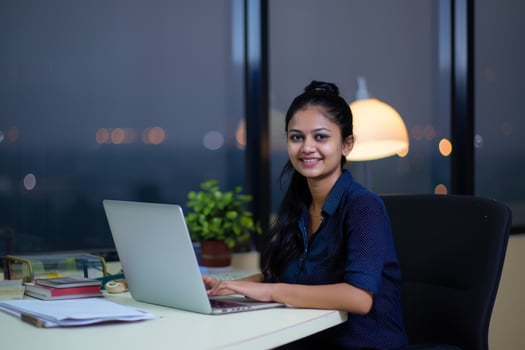 A young indian woman sitting at her desk working on laptop and smiling into the camera photo