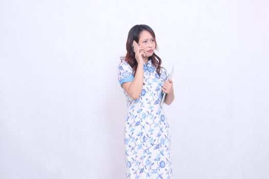 beautiful Indonesian Chinese woman looking obliquely at top left wearing modern Chinese blue dress holding tablet while making phone call isolated on white background photo
