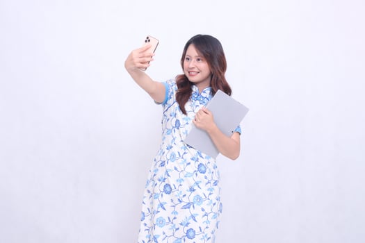 beautiful asian woman wearing modern chinese blue dress obliquely cheerful holding tablet and photo looking at cellphone isolated white background
