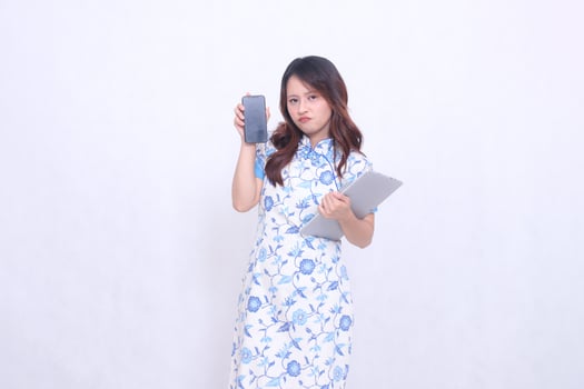 beautiful asian woman wearing blue chinese dress upset holding tablet and looking at cell phone screen isolated white background photo