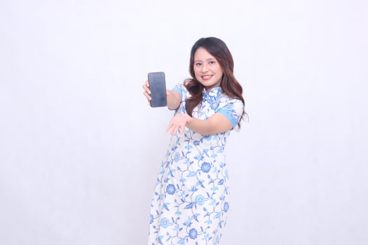 young Indonesian Javanese girl wearing a blue traditional Chinese dress cheerfully holding a gadget showing her cellphone screen raised to the front isolated on a white background photo