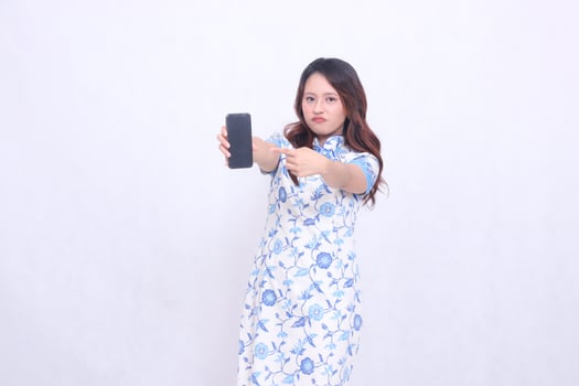 young Indonesian Javanese woman wearing blue traditional Chinese dress frowning holding gadget wide showing cell phone screen forward pointing to the side isolated white background photo