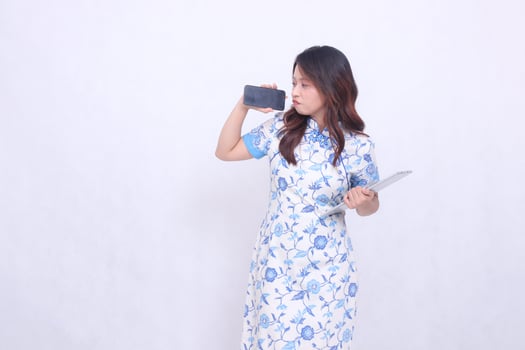 beautiful asian woman wearing modern chinese blue dress looking at cell phone frowning holding tablet and looking at cell phone screen isolated white background photo