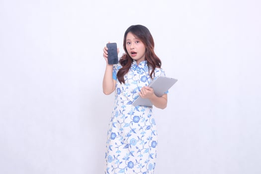Beautiful Asian woman wearing blue Chinese dress holding tablet and looking at cellphone screen isolated on white background photo