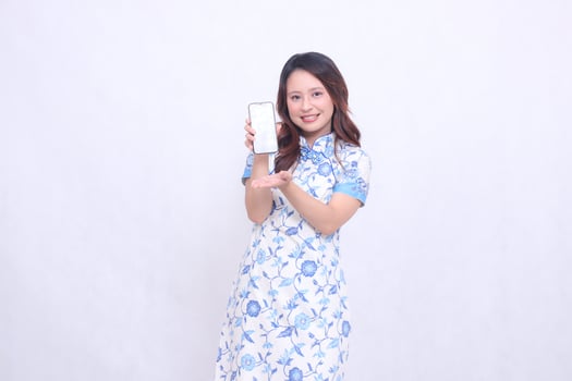Young Asian Javanese Chinese girl wearing traditional Chinese blue dress smiles promoting gadget on cell phone screen isolated white background photo