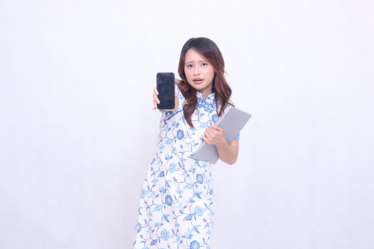 beautiful asian woman wearing blue chinese dress looking at cell phone shocked holding tablet and looking at cell phone screen straight isolated white background photo