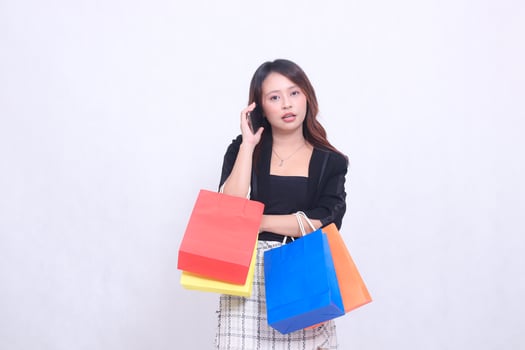 Beautiful Asian woman customer 20s formal office shocked confused while on the phone with arms crossed while carrying shopping paper bag isolated white background photo