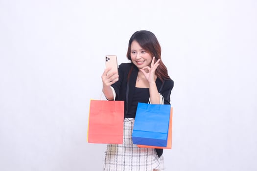 beautiful asian woman customer 20s formal office cheerful smile looking at cell phone hand gesture okay while carrying shopping paper bag isolated white background photo