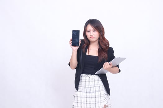 Beautiful Indonesian woman in her 20s formal office frowning looking at a black cellphone gadget and carrying a tablet for promo isolated on a white background photo