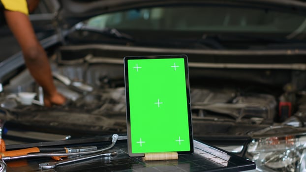 Close up shot of tablet placed on working bench in busy garage next to professional tools while engineer fixes vehicle. Mockup device in repair shop with precise expert working in blurry background photo