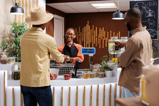 In grocery store, African American female vendor assists clients, promoting sustainability with fresh products in bio packaging. At counter, black woman taking items from male customer for weighing. photo
